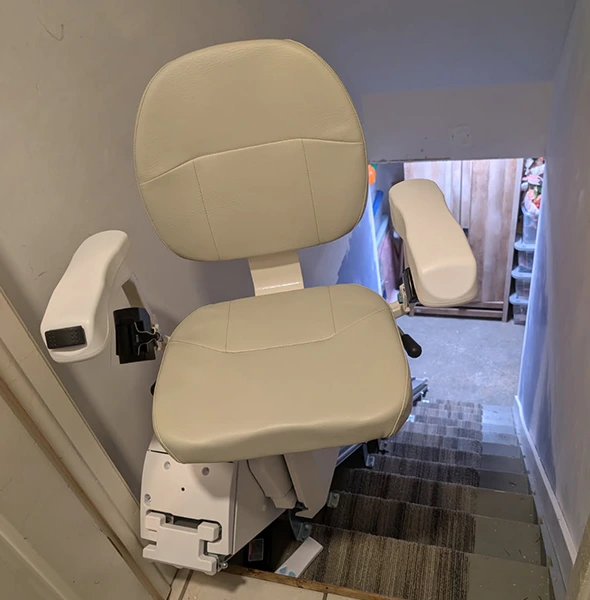 Cream-colored stair lift chair mounted on a wall beside a carpeted staircase.